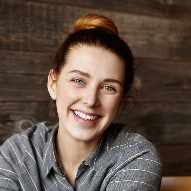 Headshot of cute girl with hair bun spending lunch break at c restaurant with wooden walls, sitting alone and waiting for her friends. Happy cheerful Caucasian woman with joyful smile posing indoors