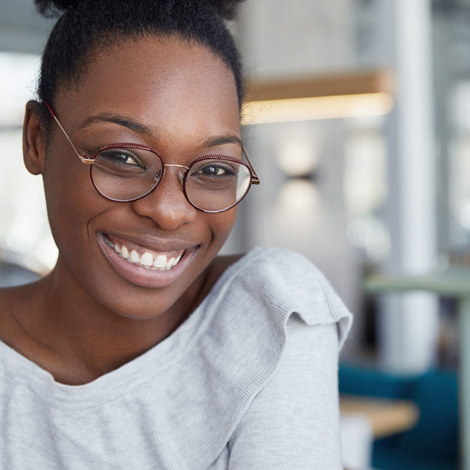 Headshot of positive attractive dark skinned African female in round glasses, expresses pleasant emotions, feels happy after recieving praise from boss for successfully done work or project.